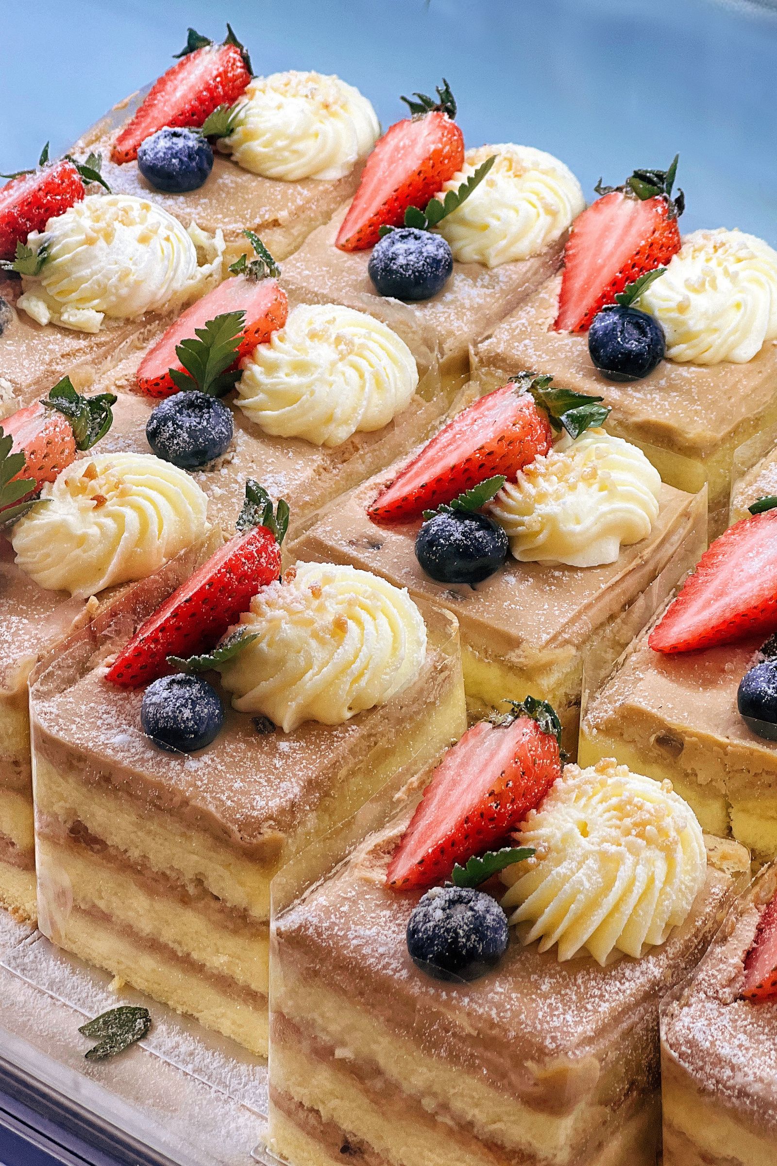 Rows of layered cream cake slices with a piped cream rosette, a strawberry half, a blueberry and a mint leaf, dusted with icing sugar.