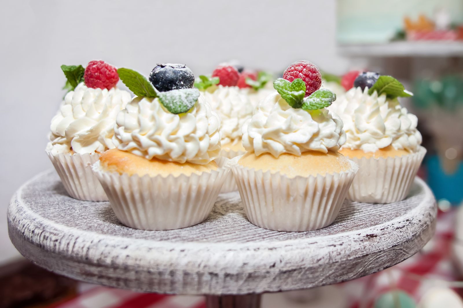 Four cupcakes with piped white cream, topped with raspberries, blueberries and mint leaves, on a distressed-white wooden round stand.