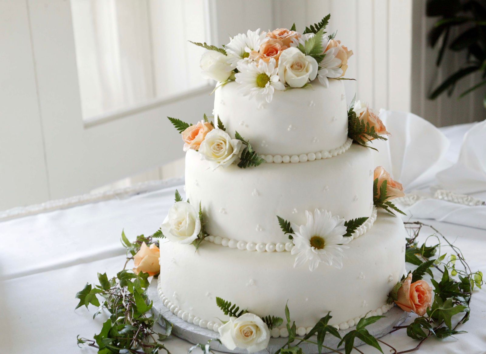 A three-tier white-iced cake with piped bead borders, decorated with white roses, peach roses, daisies, ferns and ivy, on a white-draped table by a window.